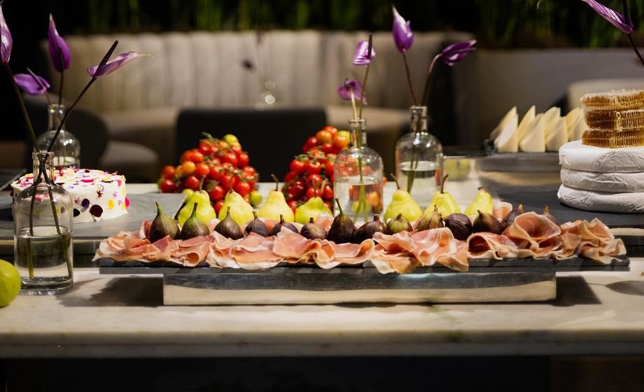 Mesa de buffet com presunto cru, figos, peras, tomates-cereja e queijos, decorada com flores roxas em vasos de vidro