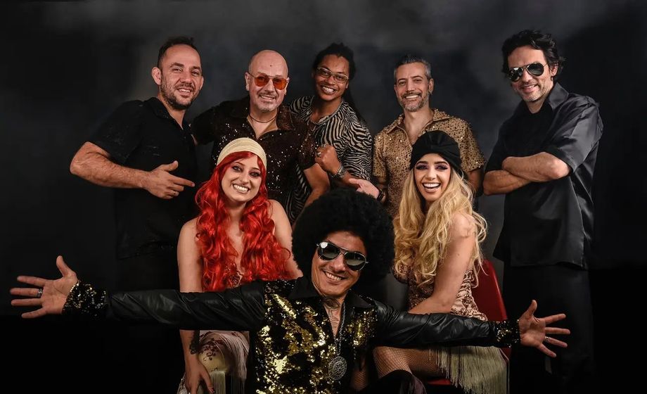 Integrantes da Banda Nega Fulô posam em figurinos brilhantes de festa disco; à frente, o vocalista de cabelo black power e óculos escuros abre os braços